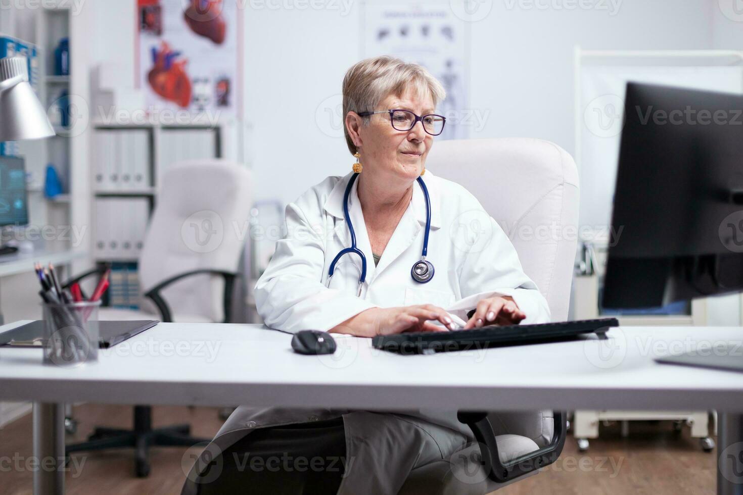 Sénior femelle médecin dans laboratoire manteau, des lunettes et stéthoscope plus de sa cou dactylographie sur ordinateur séance dans médical Bureau en utilisant à la recherche à bureau souriant tandis que travail dans médical Bureau photo