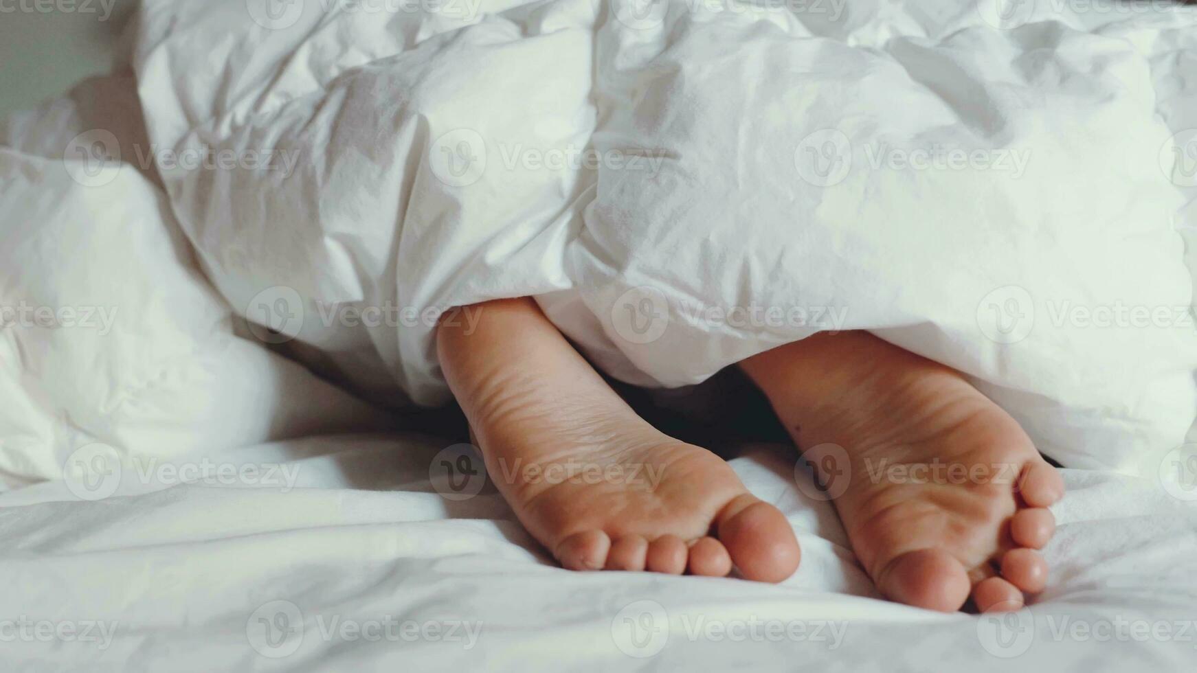 enfant pieds nus en dessous de blanc duveteux couverture. enfant cache pieds en dessous de chaud coton couverture. sommeil et relaxation dans hôtel, paresseux Matin pour les enfants. réveiller en haut avoir prêt pour école. photo