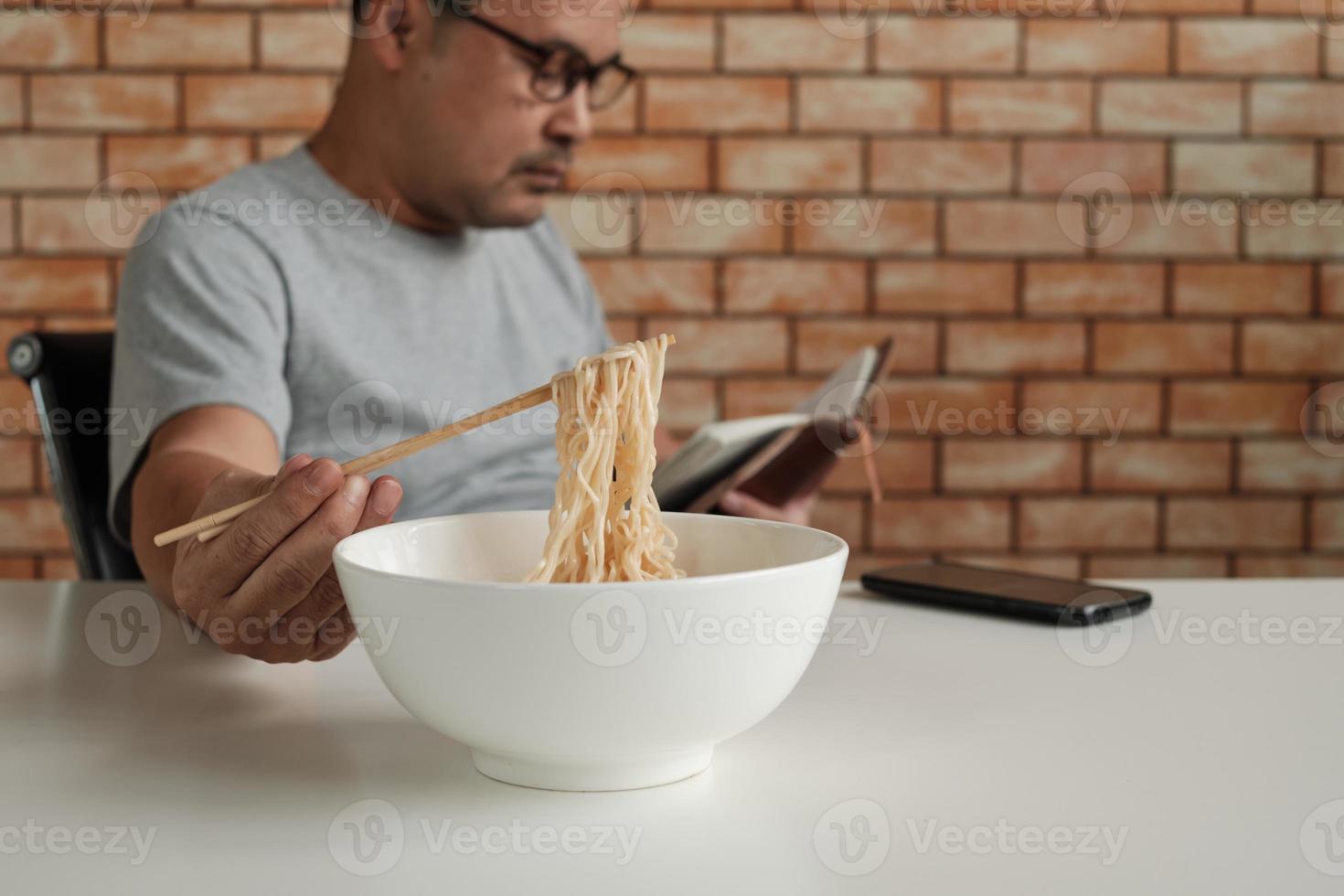 un travailleur asiatique lit un carnet de rendez-vous tout en mangeant des nouilles instantanées dans un bol blanc avec des baguettes sur une table dans un bureau de fond de mur de briques pendant une pause déjeuner, un mode de vie malsain à la hâte. photo