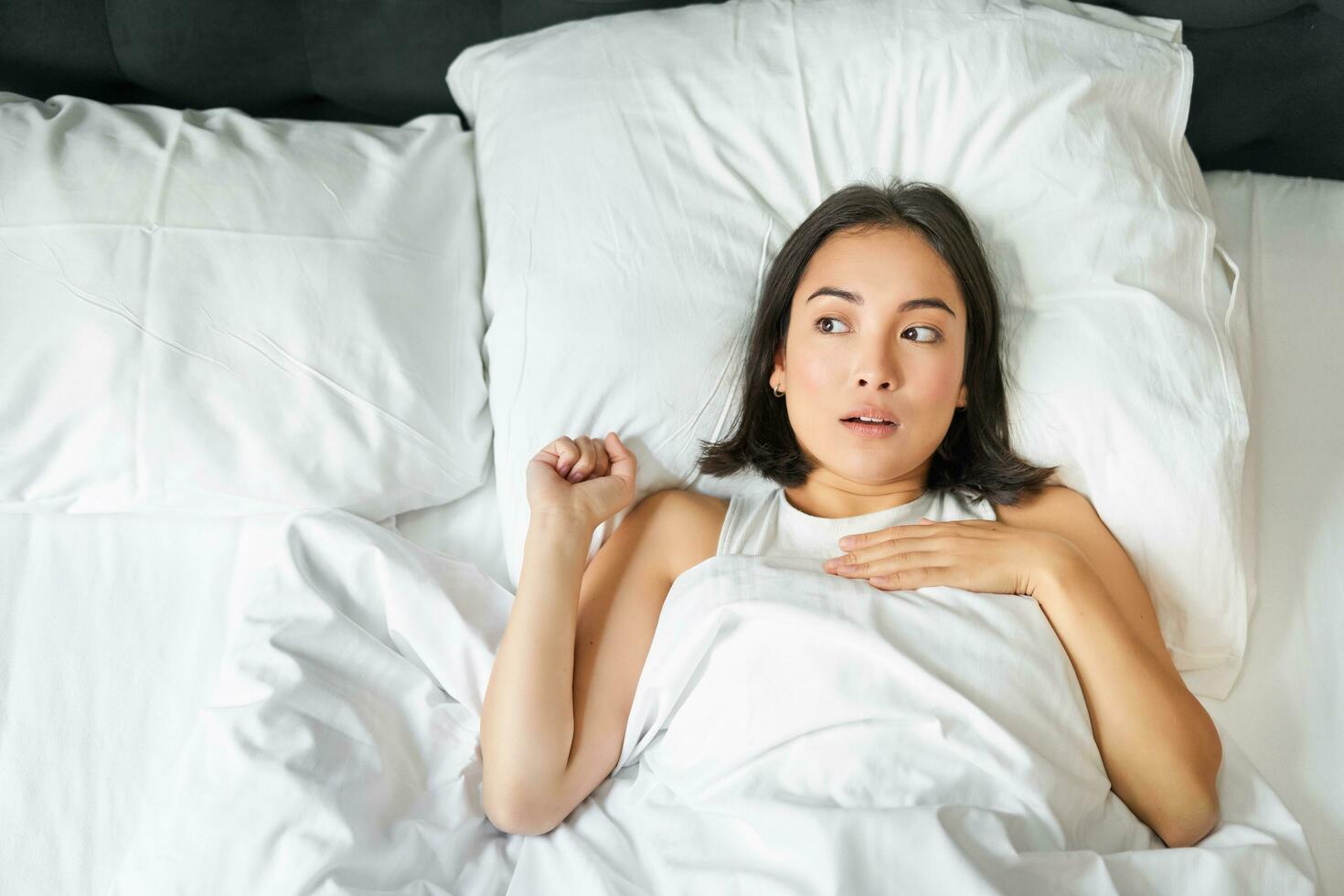 portrait de asiatique femme mensonge dans lit avec sous le choc affronter, à la recherche surpris et bouleversé, halètement de qch, rester seul dans sa chambre photo