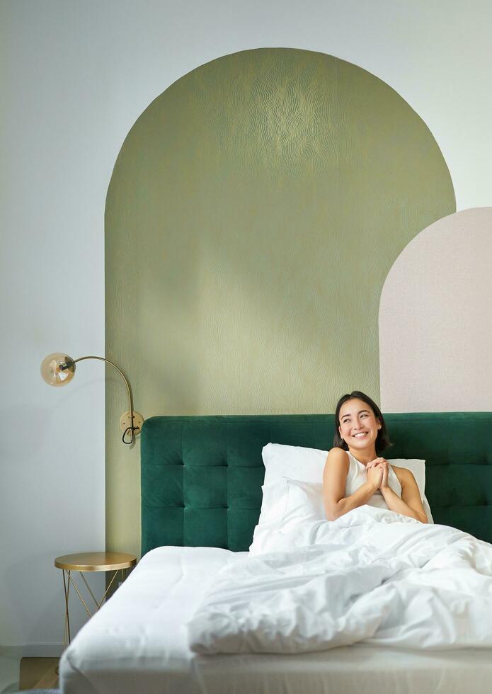 verticale coup de magnifique asiatique fille mensonge dans chambre avec sophistiqué intérieur, souriant et à la recherche content dans matin, dépenses temps dans lit photo