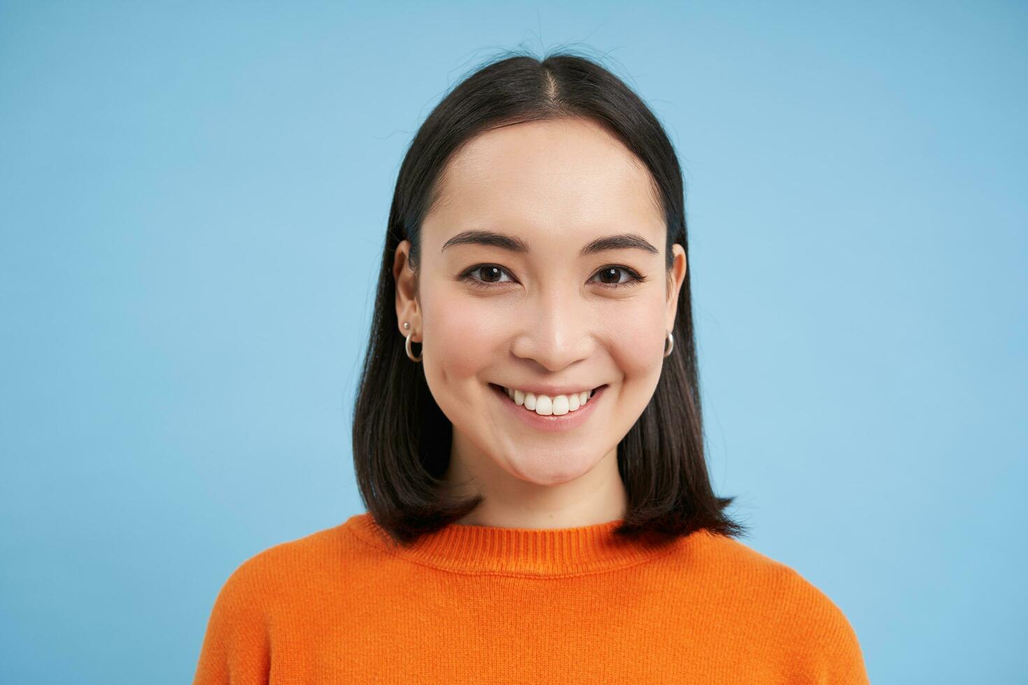 proche en haut portrait de asiatique fille avec parfait en bonne santé sourire et Naturel magnifique affronter, regards content à caméra, des stands plus de bleu Contexte photo