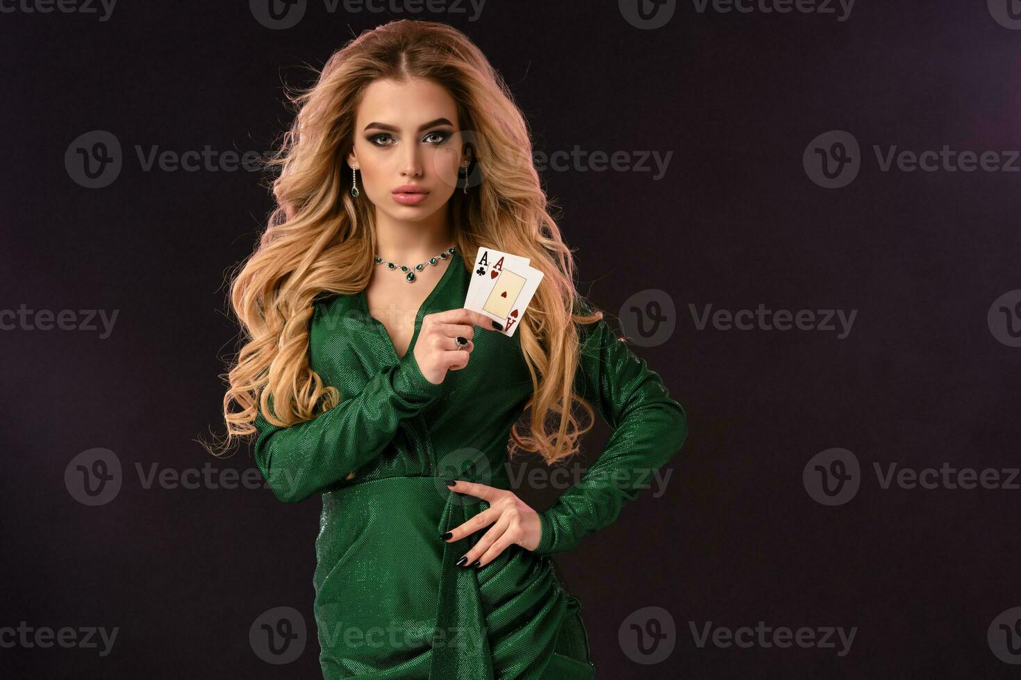blond femelle dans vert élégant robe et bijoux. mettre main sur hanche, montrant deux en jouant cartes, posant sur noir Contexte. poker, casino. fermer photo