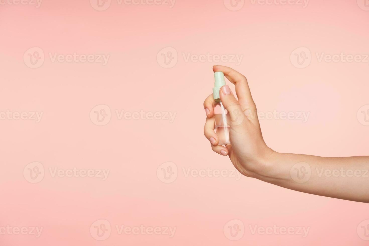 côté vue de transparent vaporisateur bouteille avec liquide étant tenue par Jeune féminin main avec nu manucure tandis que Aller à pousser bouton, isolé plus de rose Contexte photo