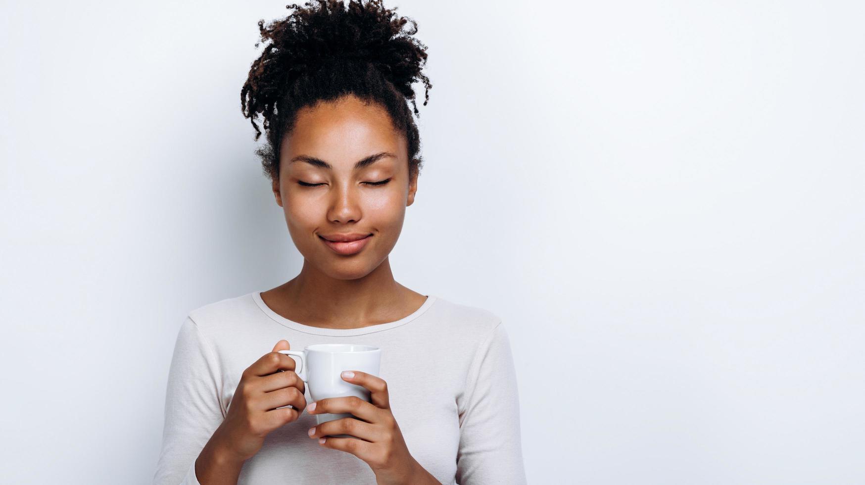 belle fille afro-américaine tenant une tasse bio, fermant les yeux en sirotant un verre sur fond blanc photo