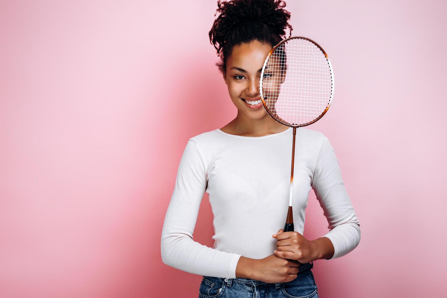 belle fille souriante se dresse sur un fond de mur rose, se couvre le visage avec une raquette photo