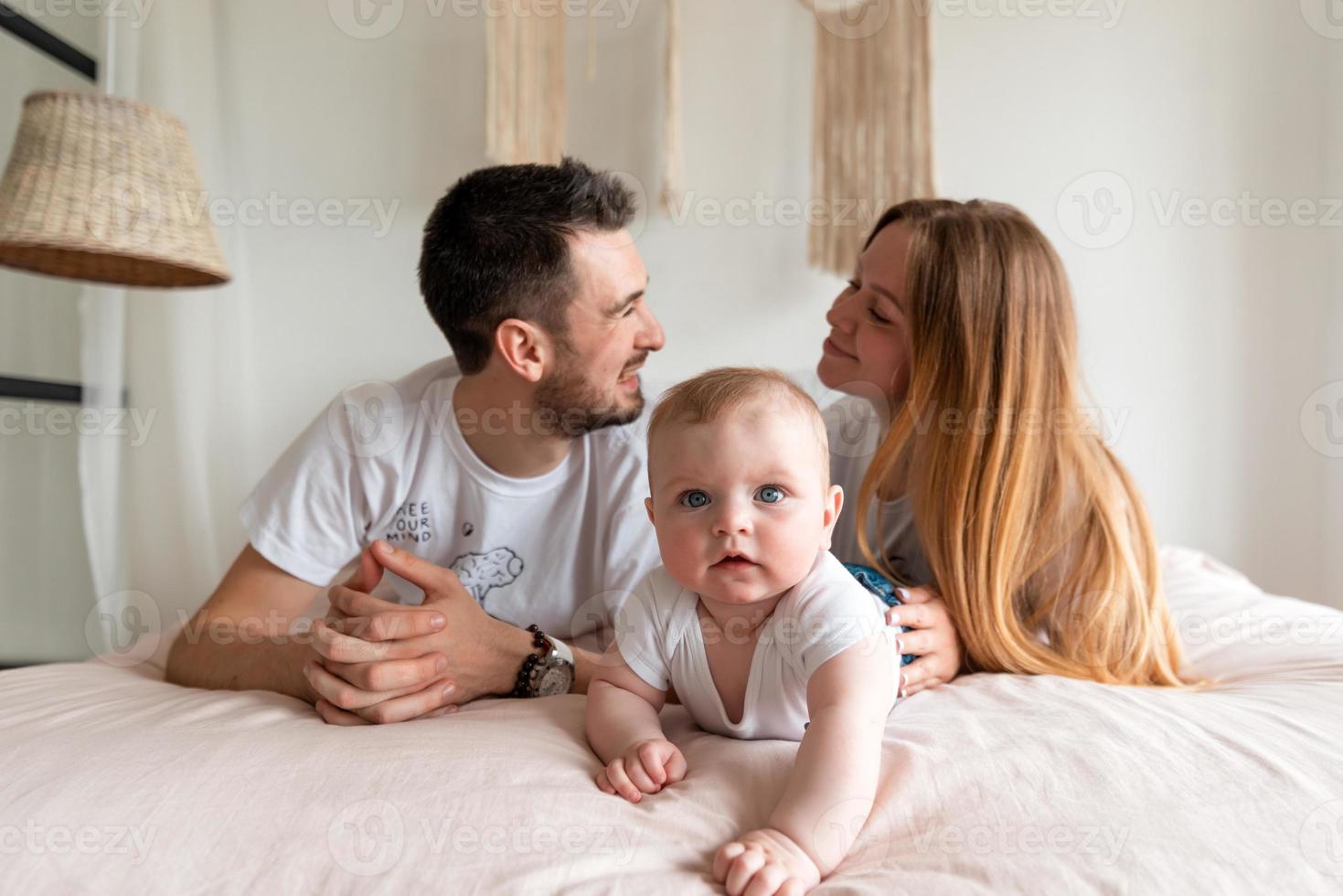 famille heureuse, mère, père et fille sur un lit blanc dans une chambre ensoleillée. les parents et un petit enfant se reposent à la maison. photo