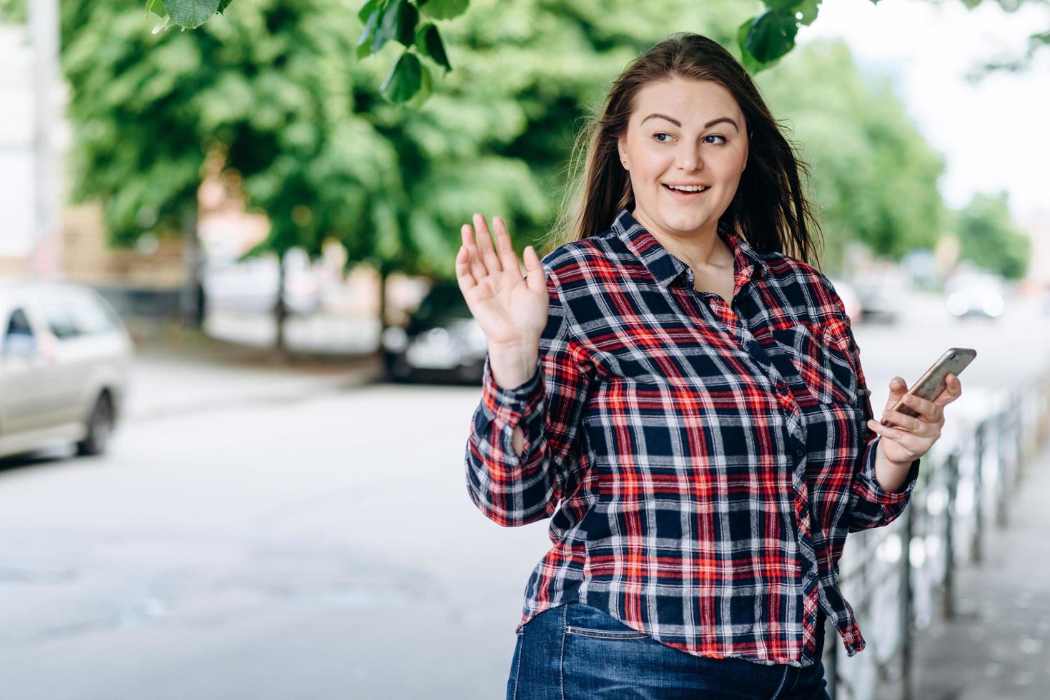 jolie fille dans la rue avec un téléphone dans les mains, souriant à quelqu'un et agitant la main photo