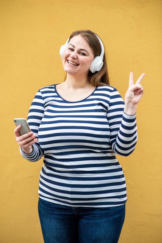jolie femme souriante dans des écouteurs sans fil blancs montre un geste de paix sur fond orange photo