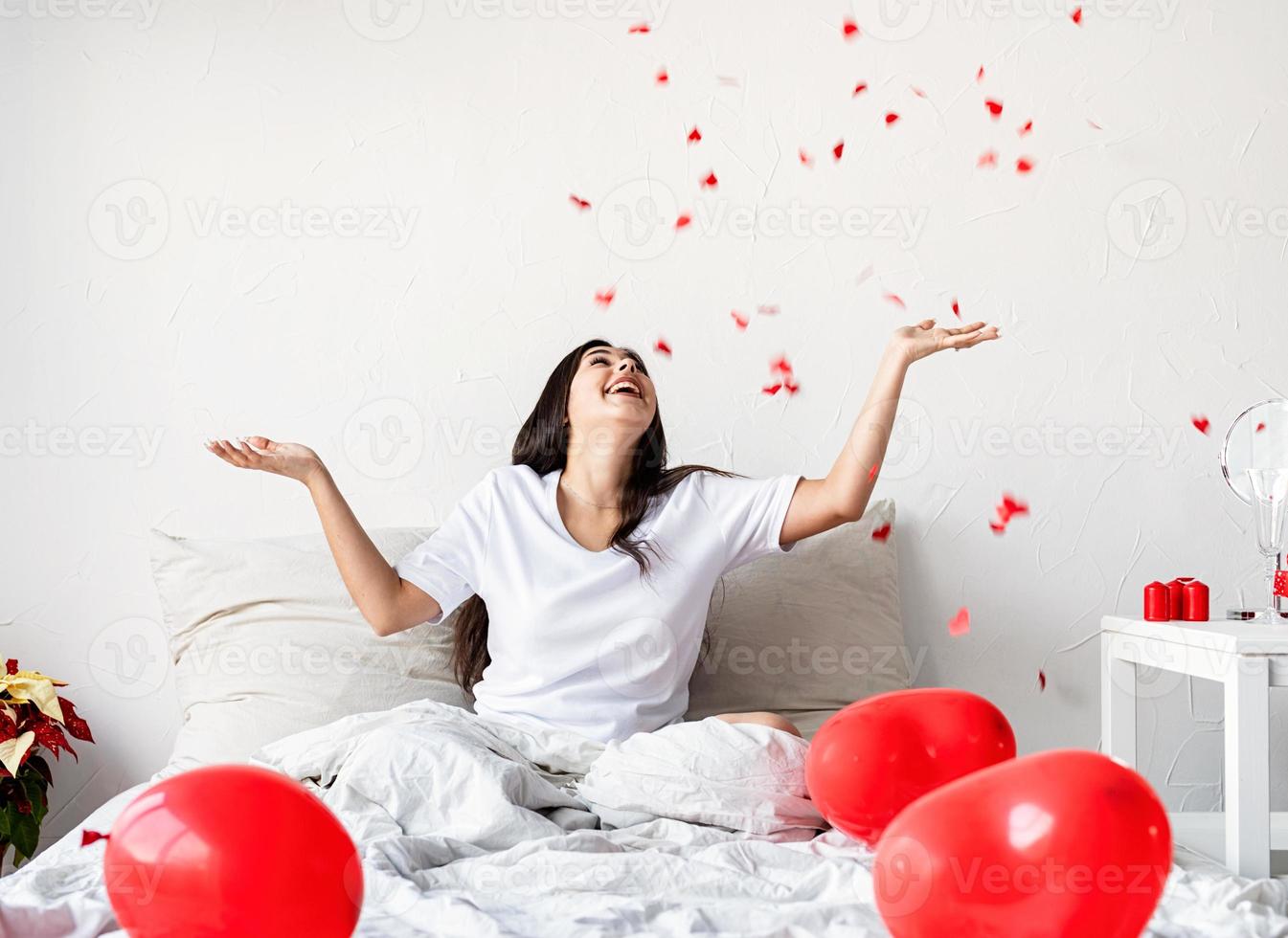Jeune femme brune heureuse assise dans le lit avec des ballons en forme de coeur rouge jetant des confettis en l'air photo
