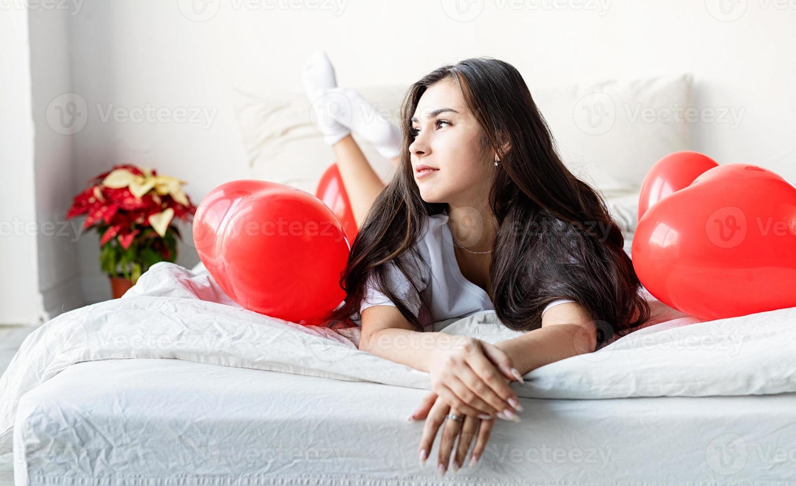 jeune femme brune heureuse pose dans le lit avec des ballons en forme de coeur rouge photo