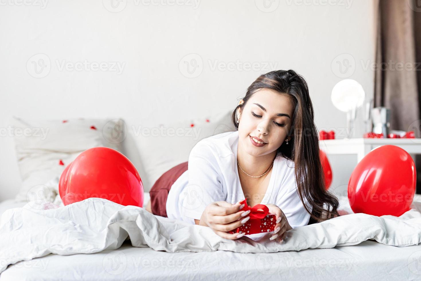 Jeune femme brune heureuse allongée dans le lit avec des ballons en forme de coeur rouge déballant le présent photo