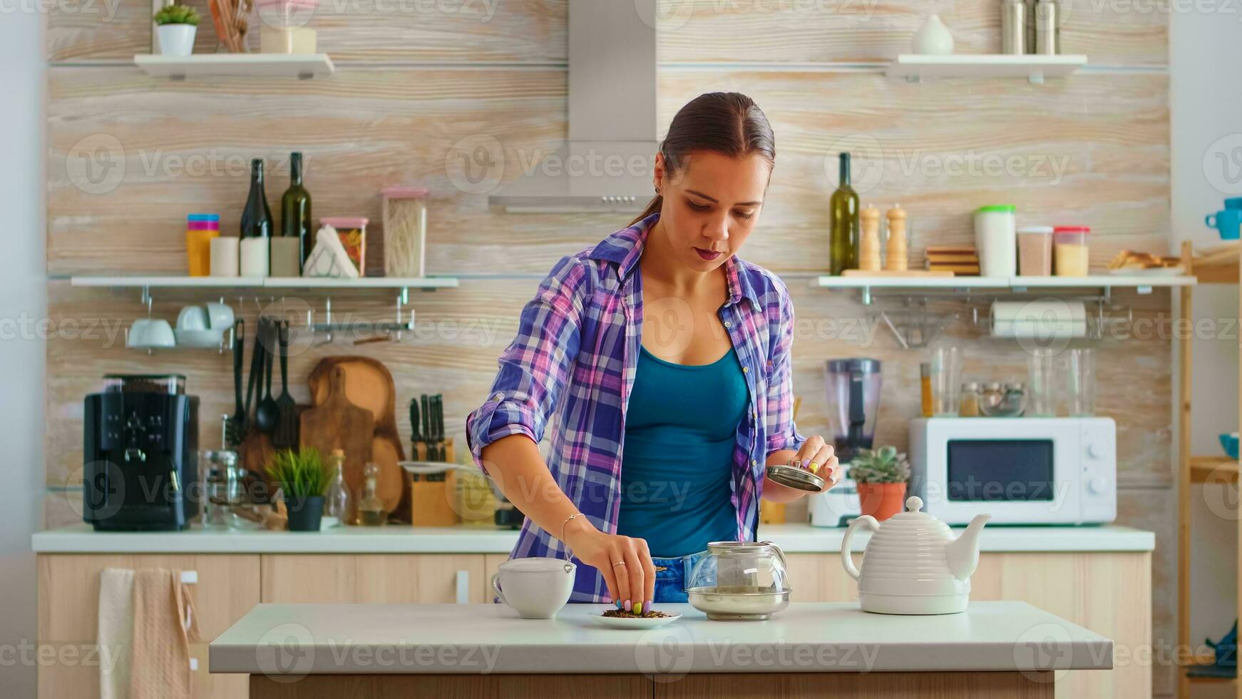 Jeune femme en train de préparer vert thé pour petit déjeuner dans une moderne cuisine en utilisant théière séance près le tableau. en mettant avec mains, pressage à base de plantes, en bonne santé, thé feuilles, dans pot, dans le Matin. photo