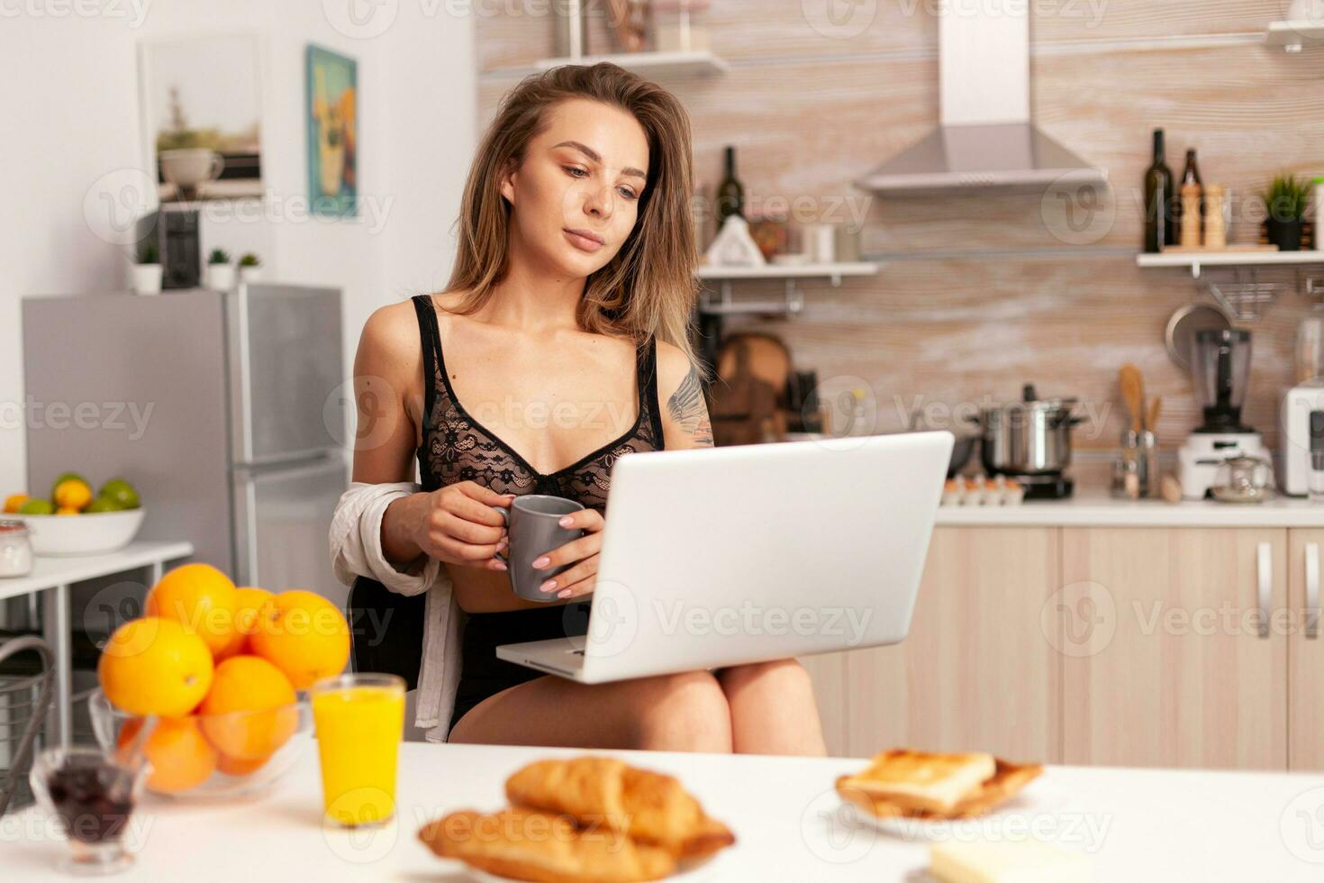 insouciant femme dans sexy lingerie en portant tasse de chaud café en utilisant portable dans cuisine. attrayant blond Dame avec tatouages dactylographie sur PC séance dans le cuisine habillé dans séduisant sous-vêtements souriant photo