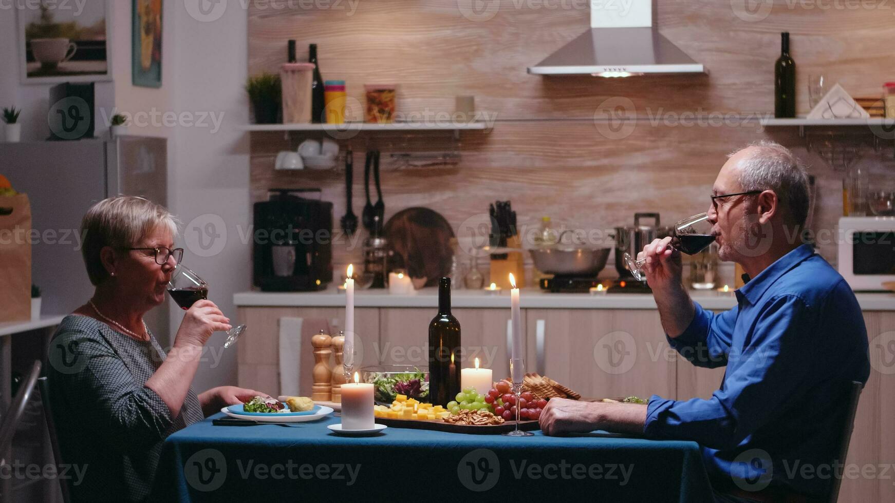 personnes âgées mature couple en buvant une verre de du vin pendant romantique dîner. Sénior vieux gens tintement, séance à le table dans cuisine, profiter le repas, célébrer leur anniversaire dans le à manger chambre. photo