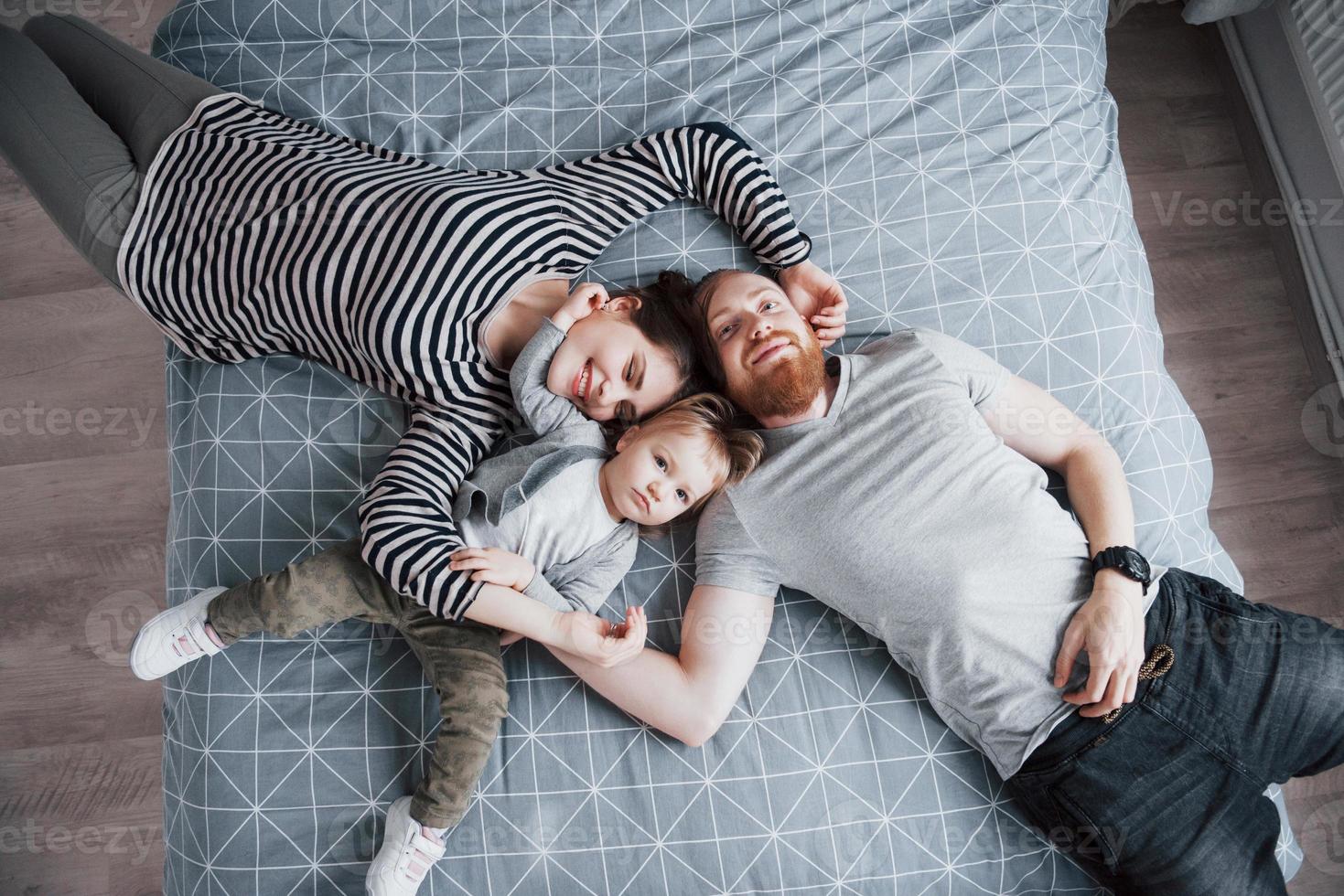 vue de dessus de la belle jeune mère, du père et de leur fille regardant la caméra et souriant en position couchée sur le lit tête à tête photo