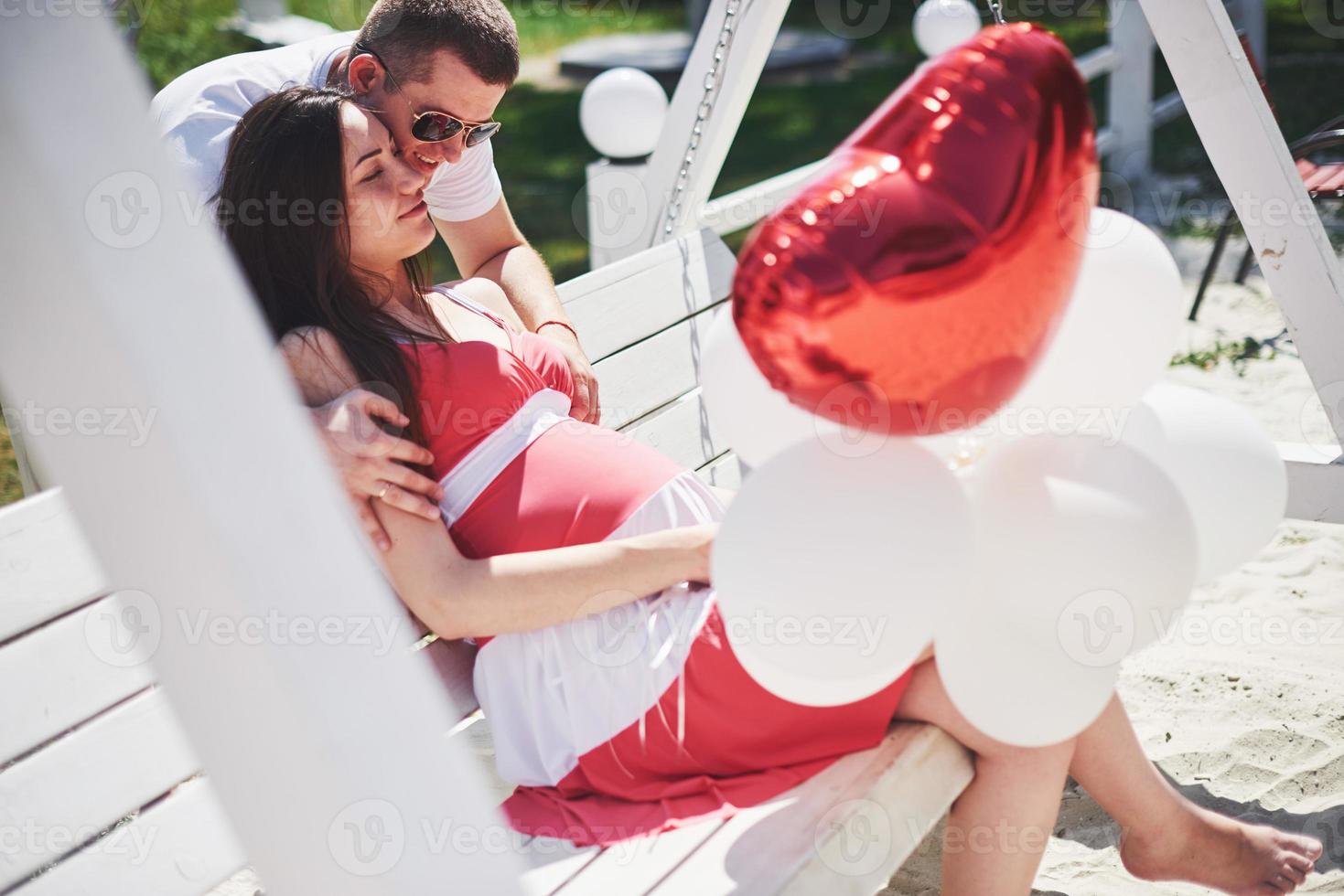 bébé en attente. femme enceinte avec son mari bien-aimé s'asseoir sur un banc. la main du mari embrasse une femme au ventre rond. parentalité. femme enceinte vêtue d'une robe blanche en cerise avec un noeud rouge. fermer. neuf mois photo