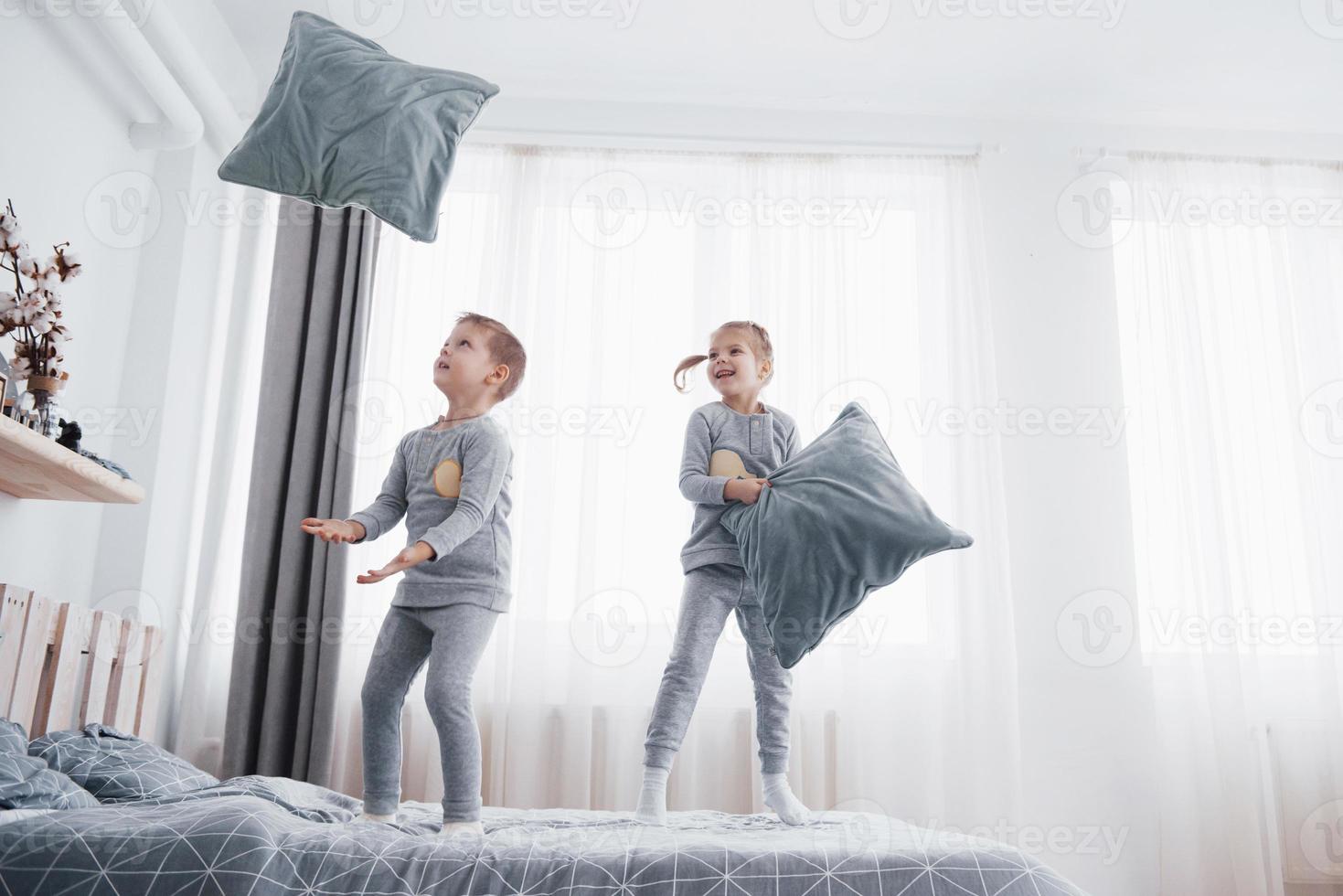 enfants jouant dans le lit des parents. les enfants se réveillent dans une chambre blanche ensoleillée. garçon et fille jouent en pyjama assorti. vêtements de nuit et literie pour enfant et bébé. intérieur de pépinière pour enfant en bas âge. matin en famille photo