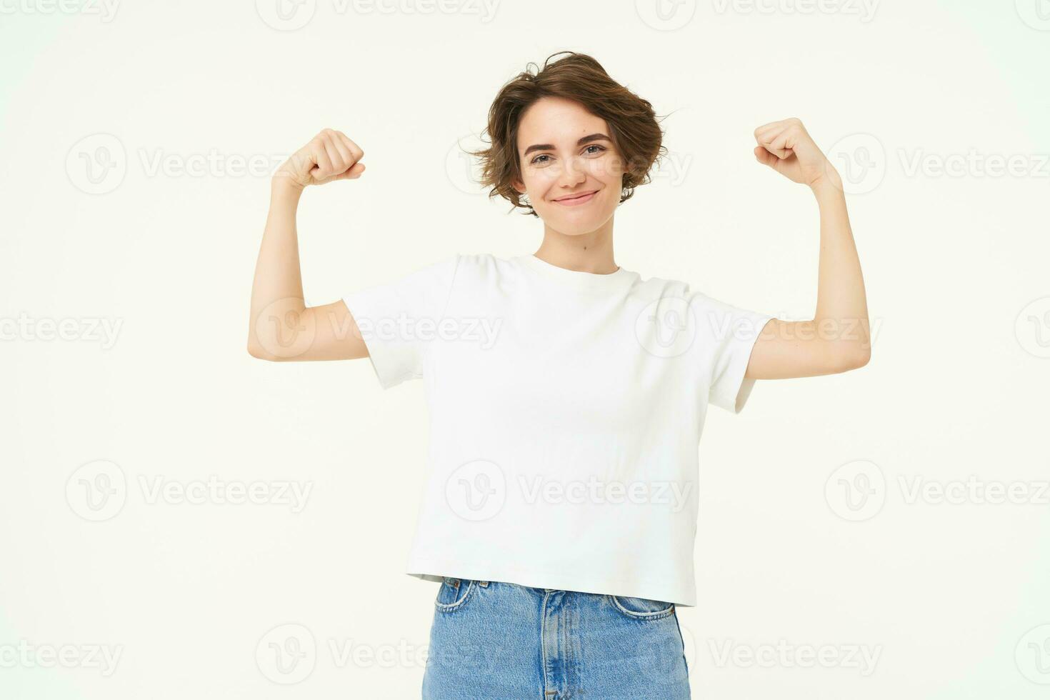 portrait de sur de soi et fort Jeune femme, fléchissement biceps, spectacles sa muscles sur bras avec fier souriant affronter, posant plus de blanc Contexte photo