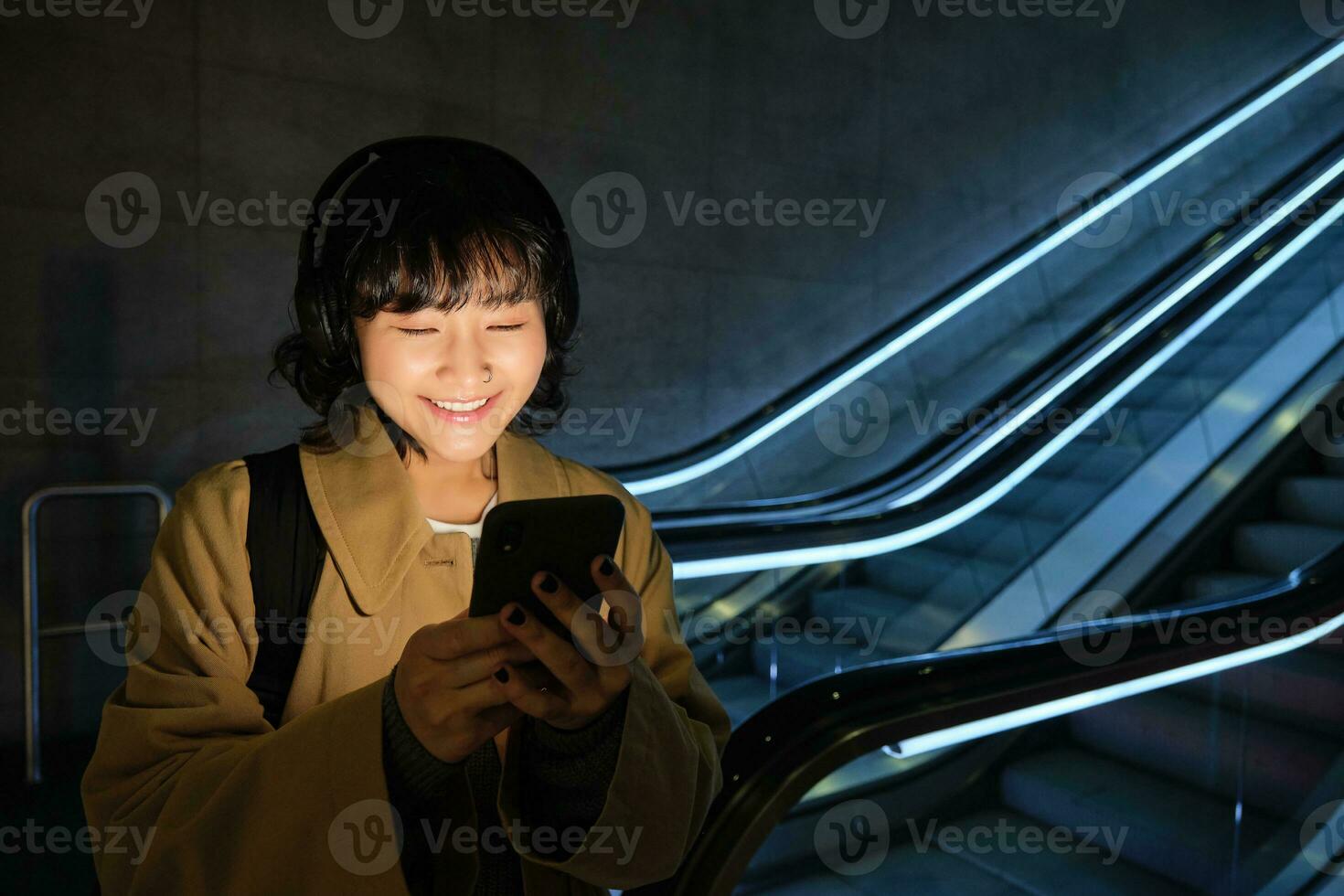 content asiatique femme dans métro, permanent près escalier mécanique, en portant téléphone intelligent et écoute la musique dans écouteurs, souriant heureux photo