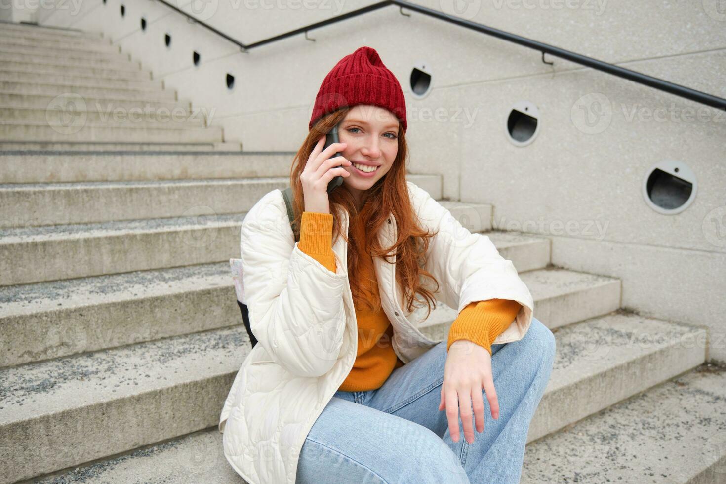 Jeune élégant roux fille dans rouge chapeau, est assis sur rue et pourparlers sur mobile téléphone, a Téléphone conversation, anneaux sa ami tandis que se détend en plein air photo