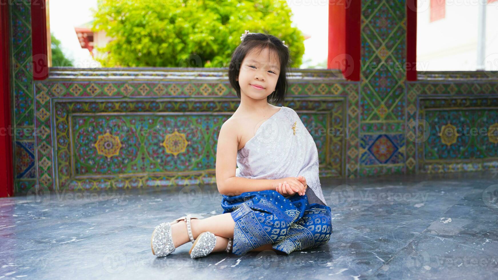 mignonne fille porte thaïlandais robe avec argent ceinture et bleu pagne taillé avec or. asiatique enfant visites temple dans Thaïlande à voir art et culture avec sa famille. enfant squats asseoir sur le sol et sourit. photo