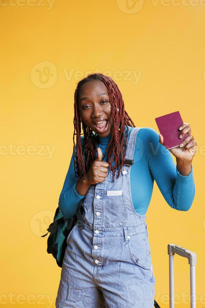 la personne avec passeport donnant les pouces en haut dans studio, profiter vacances vacances sur international destination. Jeune adulte montrant comme geste pour id passer, joyeux femme avec chariot Sacs. photo