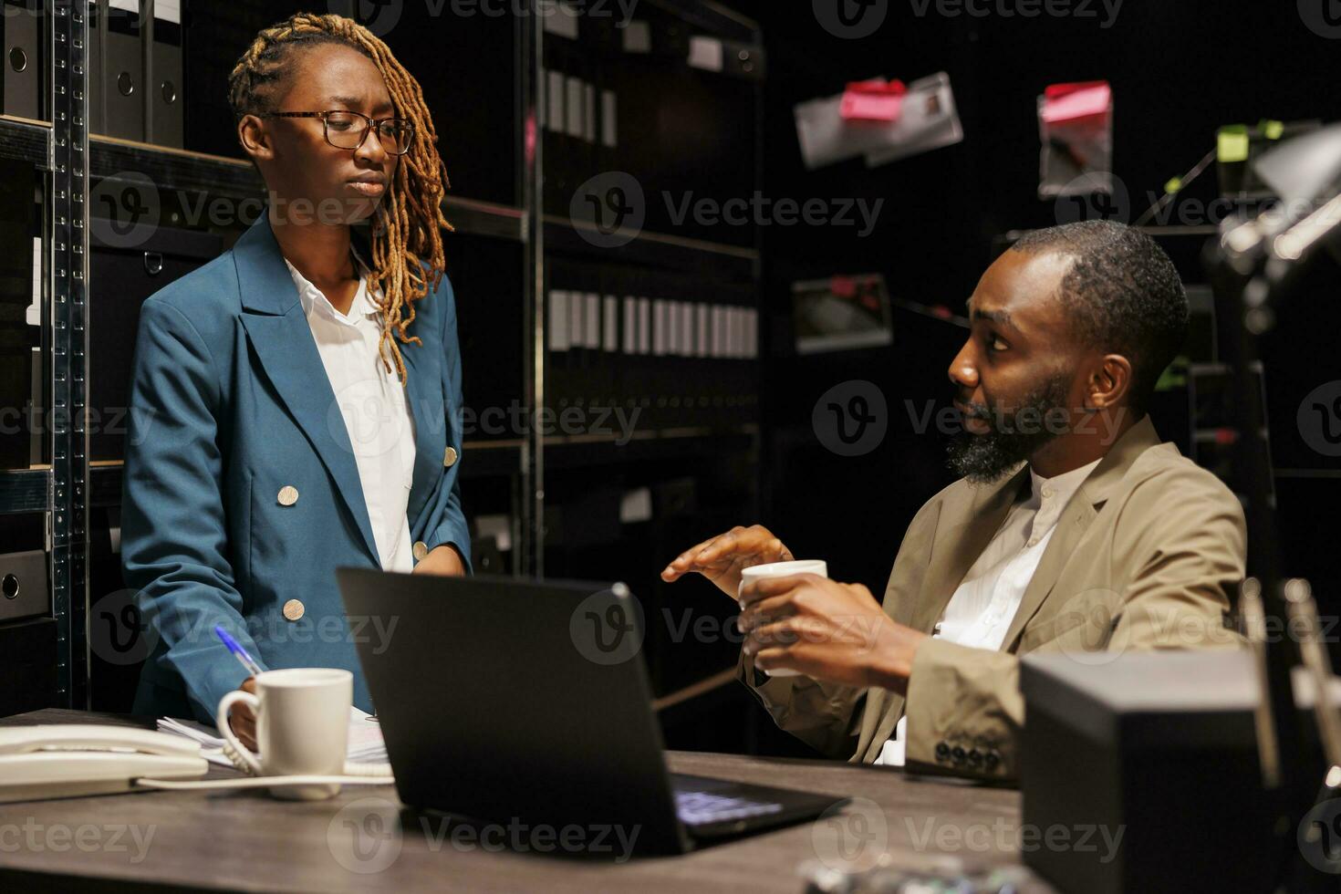 africain américain flics équipe travail dans archiver à résoudre la criminalité cas. homme détective séance à bureau près étagère plein de Dossiers et des dossiers, discuter perspicacité avec femme partenaire à nuit temps photo