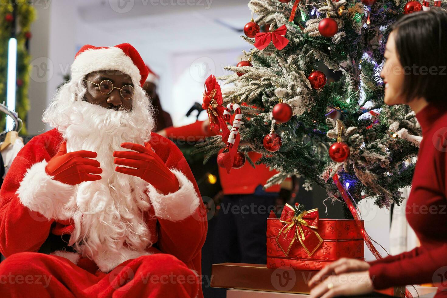 centre commercial ouvrier Père Noël claus séance suivant à Noël arbre avec asiatique client dans de fête décoré vente au détail magasin. mode boutique employé offre cadeau à content client, discuter à propos hiver vœux. photo