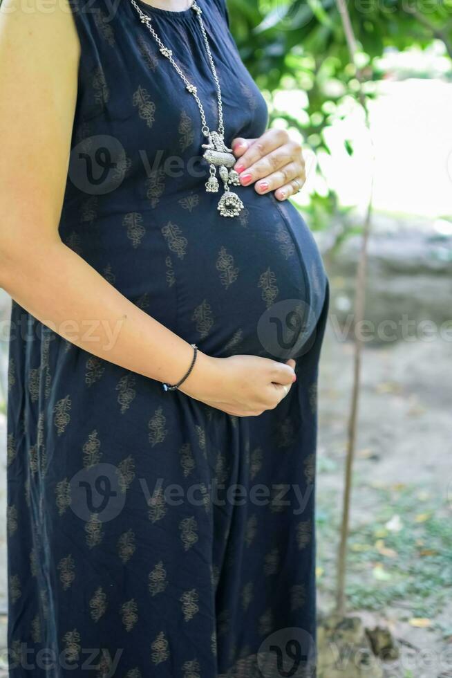 une Enceinte Indien Dame pose pour Extérieur grossesse tirer et mains sur ventre, Indien Enceinte femme met sa main sur sa estomac avec une maternité robe à société parc, Enceinte à l'extérieur maternité tirer photo