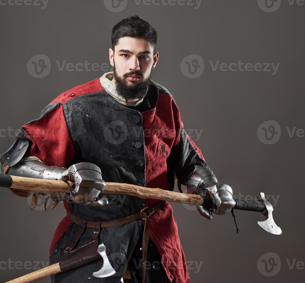 chevalier médiéval sur fond gris. portrait d'un guerrier brutal au visage sale avec une armure de cotte de mailles, des vêtements rouges et noirs et une hache de combat photo