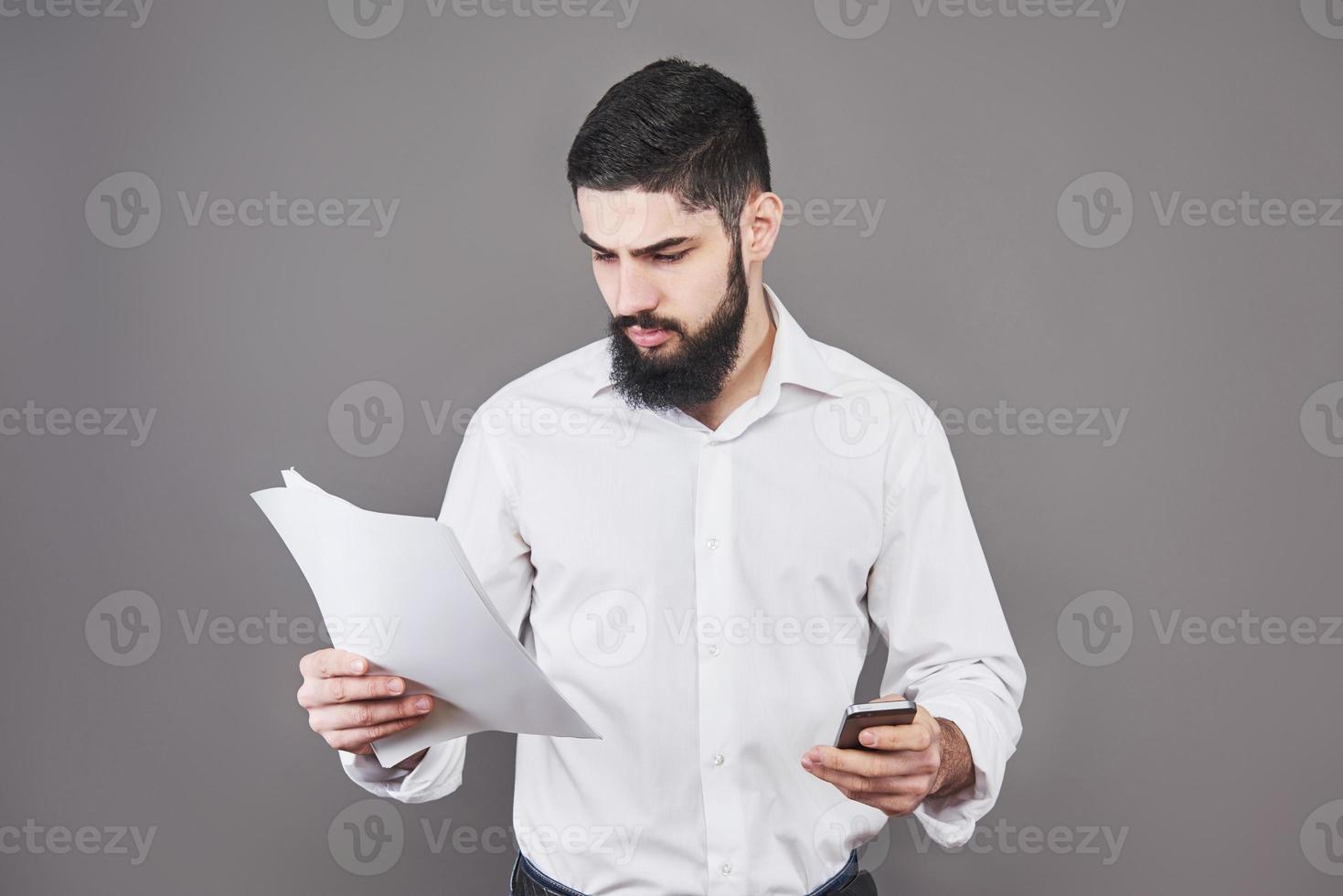 homme d'affaires avec barbe en chemise blanche tenant des documents et téléphone sur fond gris photo