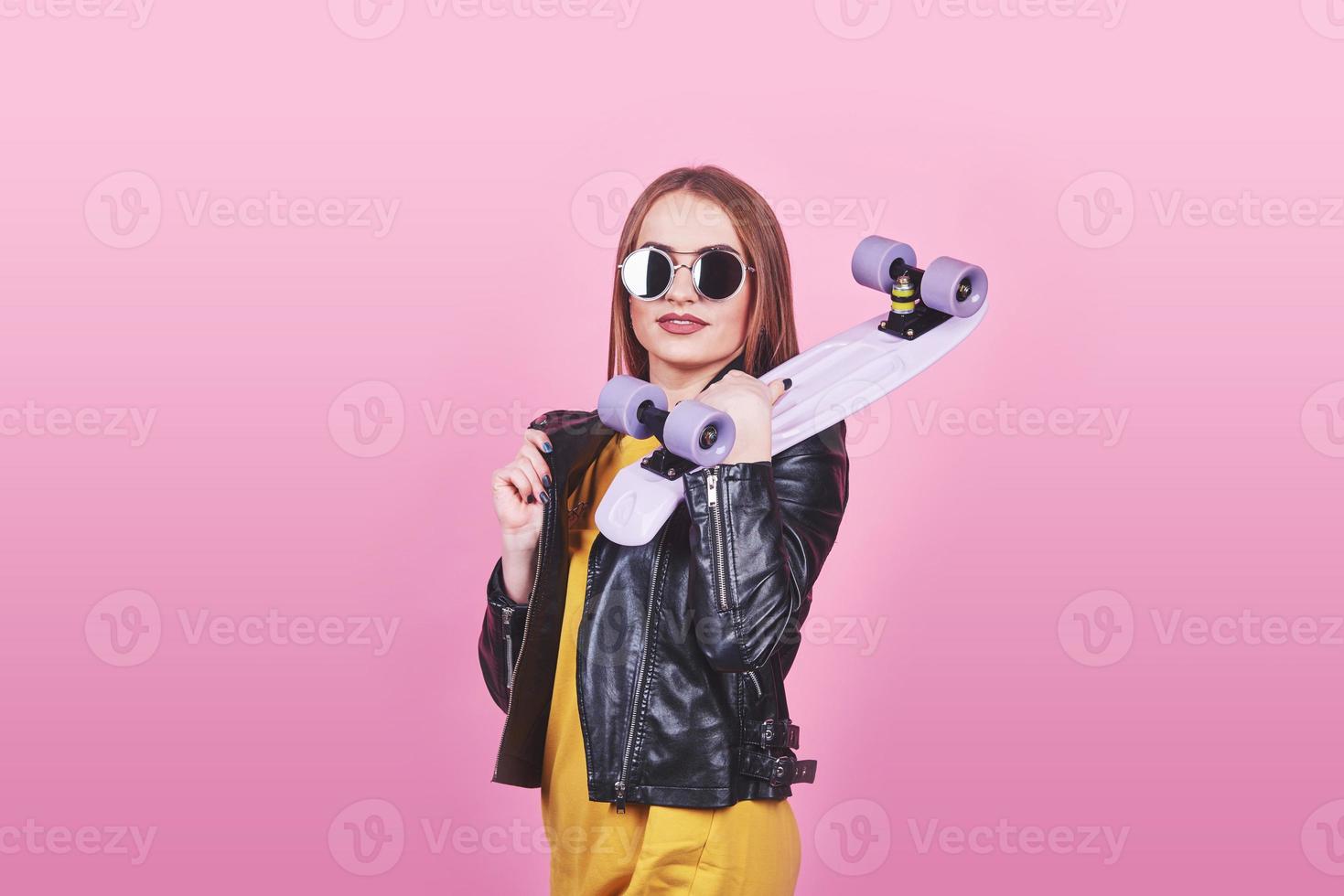 jolie fille bronzée avec le sourire au visage porte une veste en cuir noir posant avec plaisir. Photo d'intérieur d'une jolie femme à lunettes de soleil debout avec une planche à roulettes sur fond bleu