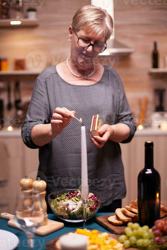 Sénior femme en utilisant allumettes à lumière bougie pour de fête dîner. personnes âgées femme attendre sa mari pour une romantique dîner. mature épouse en train de préparer de fête repas pour anniversaire fête. photo