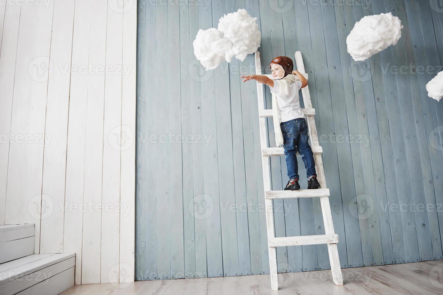 petit garçon au chapeau de pilote debout sur l'échelle dans le studio. atteindre le ciel. toucher le concept de nuages photo