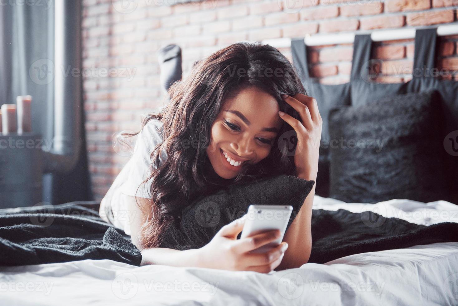 portrait d'une belle femme se réveillant dans son lit et regarde dans le téléphone. consulter les réseaux sociaux, envoyer des sms. la fille porte un t-shirt photo