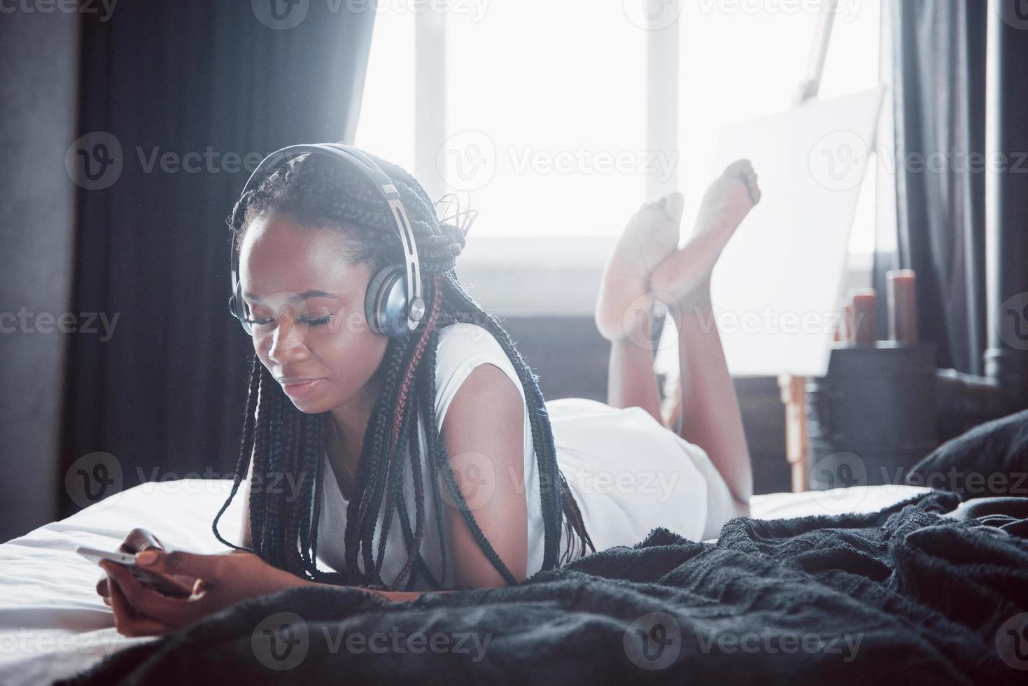 belle fille africaine en vêtements de nuit souriante écoutant de la musique dans les écouteurs allongée sur le lit photo