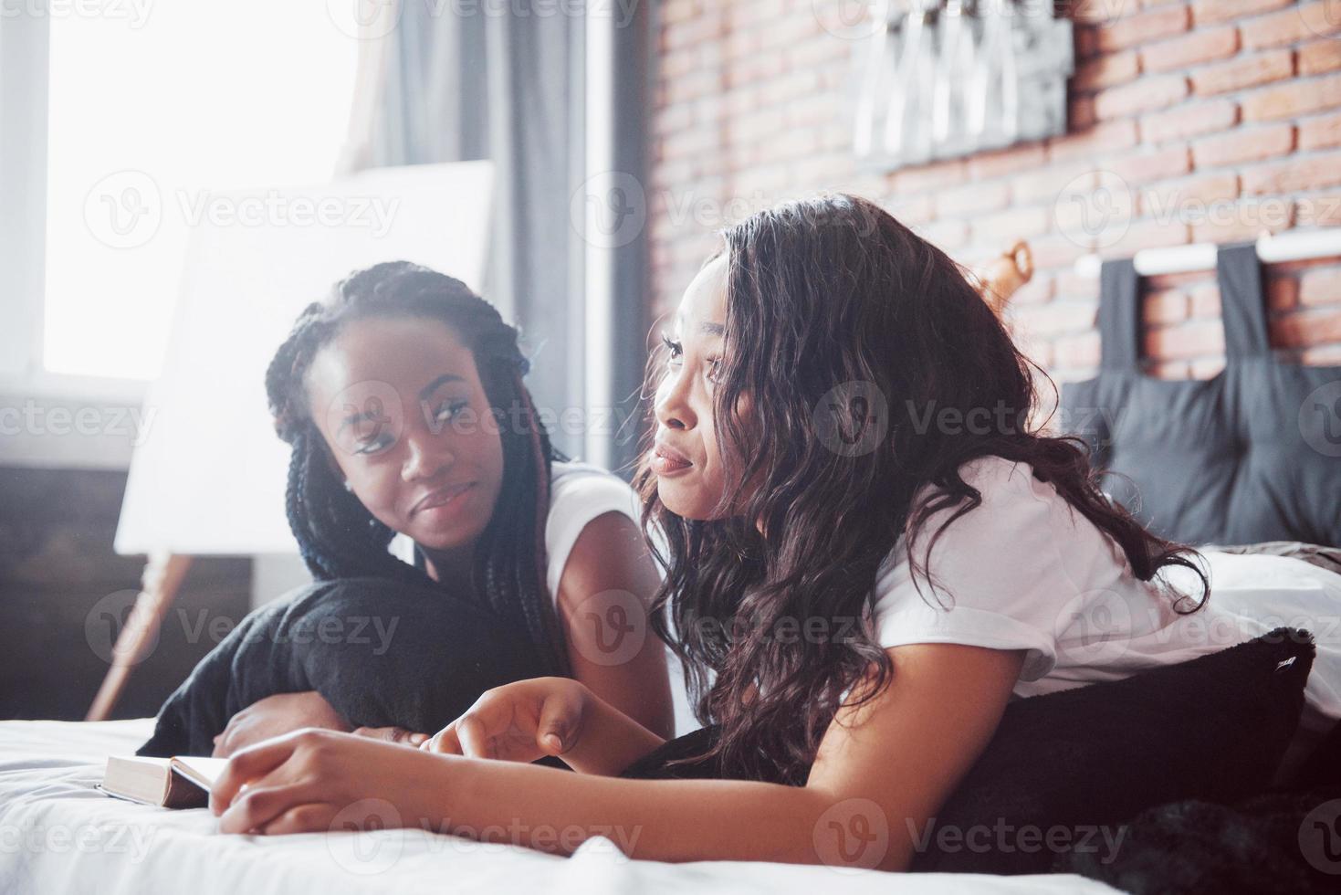 deux belles filles africaines en vêtements de nuit souriantes assises sur le lit à la maison se sont réveillées le matin par une journée ensoleillée. photo