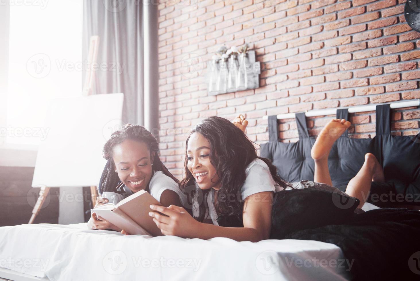 deux belles filles africaines en vêtements de nuit souriantes assises sur le lit à la maison se sont réveillées le matin par une journée ensoleillée. photo