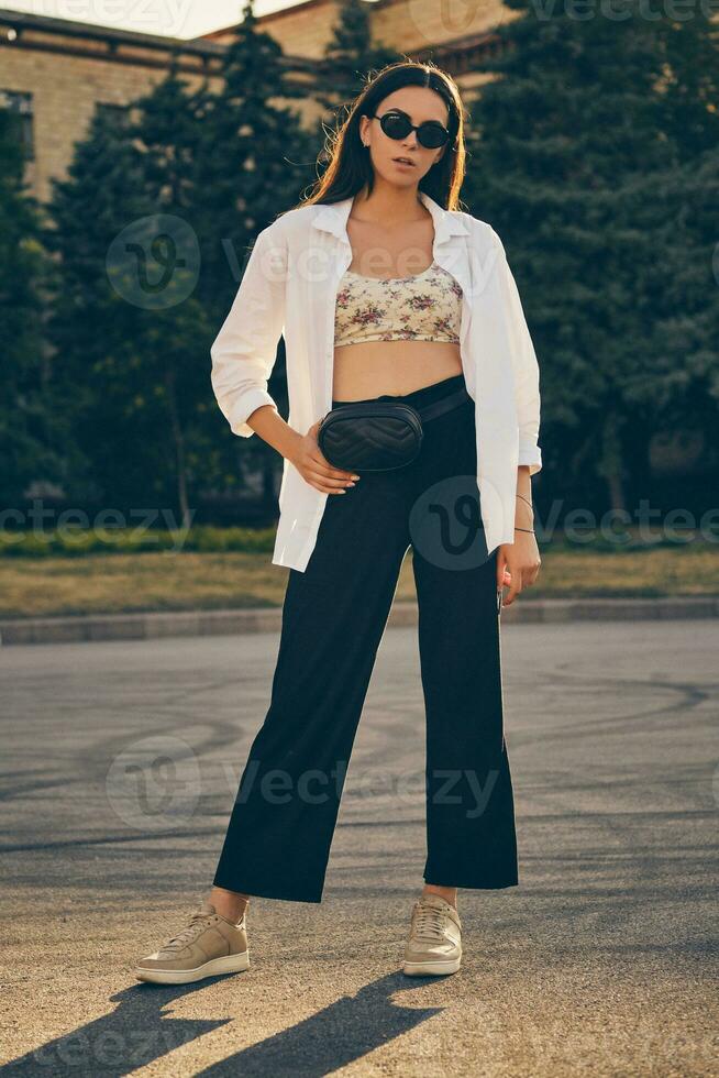 portrait de une fille dans foncé des lunettes de soleil posant dans ville. habillé dans Haut avec floral imprimer, blanc chemise, noir pantalon, taille sac et baskets. photo