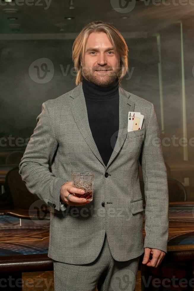 souriant homme avec deux as dans veste poche et verre de boisson dans enfumé casino photo