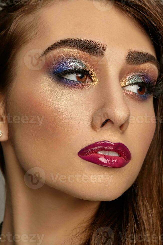 brunette femme avec luxe maquillage et parfait peau est à la recherche de côté. multicolore le fard à paupières, faux cils, brillant Bourgogne lèvres. proche en haut photo