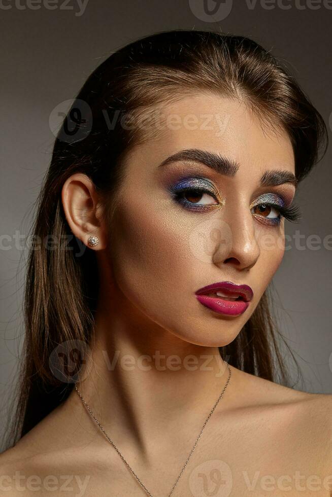 nu fille dans Collier et des boucles d'oreilles posant sur gris Contexte. luxe se maquiller, parfait peau. coloré le fard à paupières, longue cils, violet lèvres. proche en haut photo