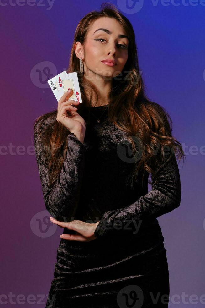 brunette femme dans noir velours robe montrant deux en jouant cartes, posant contre coloful Contexte. jeux d'argent divertissement, poker, casino. fermer. photo