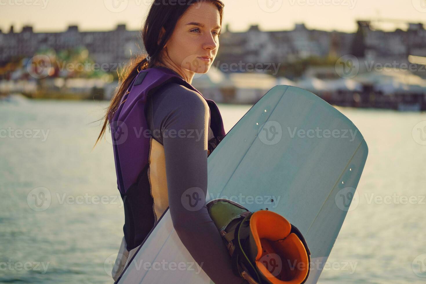 brunette fille dans une gris col roulé, spécial des sports gilet et bleu denim short est posant avec sa wakeboard sur une jetée de le côtier zone. fermer. photo