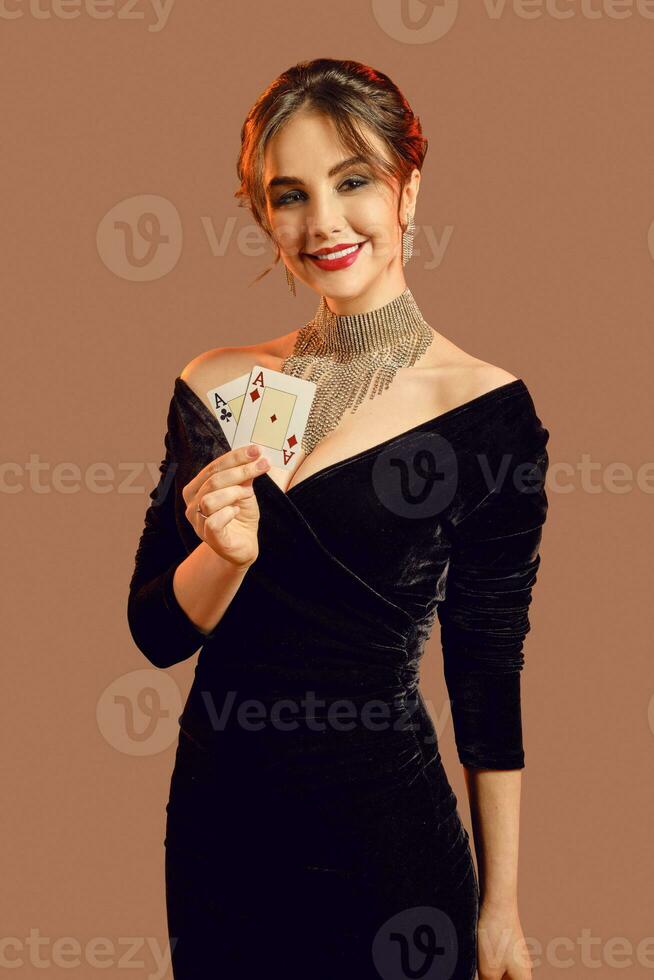 brunette femme, nu épaules, dans noir robe et bijoux. souriant, montrant deux en jouant cartes, posant sur marron Contexte. poker, casino. fermer photo