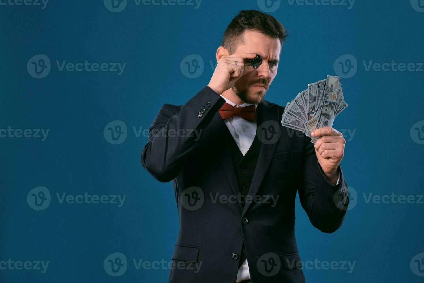 homme dans noir classique costume et rouge nœud papillon montrant un puce et certains dollar factures, posant sur bleu Contexte. jeu, poker, casino. fermer. photo