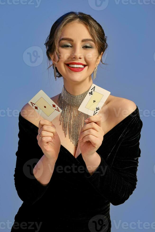 brunette modèle avec nu épaules, dans noir robe et bijoux. souriant, montrant deux en jouant cartes posant sur bleu Contexte. poker, casino. fermer photo