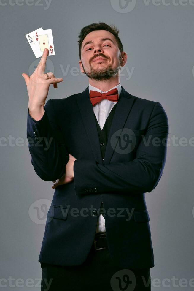 homme dans noir classique costume et rouge nœud papillon montrant deux en jouant cartes tandis que posant contre gris studio Contexte. jeu, poker, casino. fermer. photo