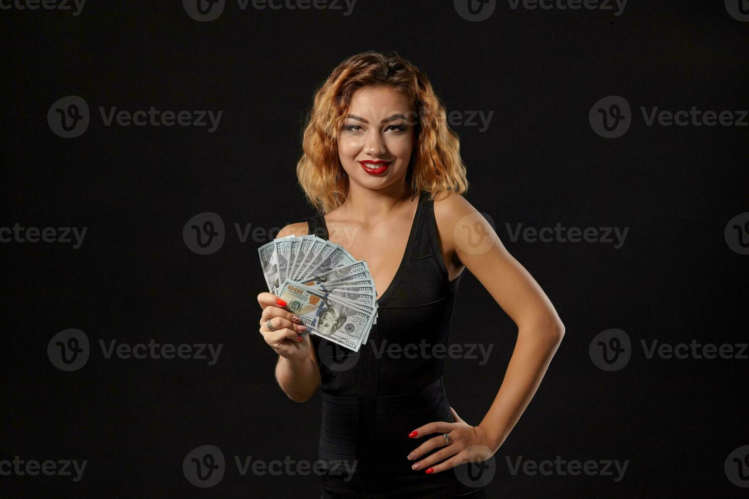 gingembre fille dans foncé robe posant en portant ventilateur de cent dollars factures dans sa mains permanent contre une noir studio Contexte. casino, poker. fermer. photo